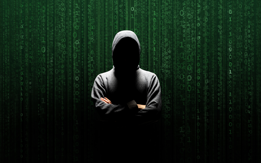Person, abgebildet als typischer "Hacker" - Hoodie der das Gesicht verdeckt. Im Hintergrund befindet sich eine grün-schwarze Wand an welcher einsen und nullen herunterrieseln.